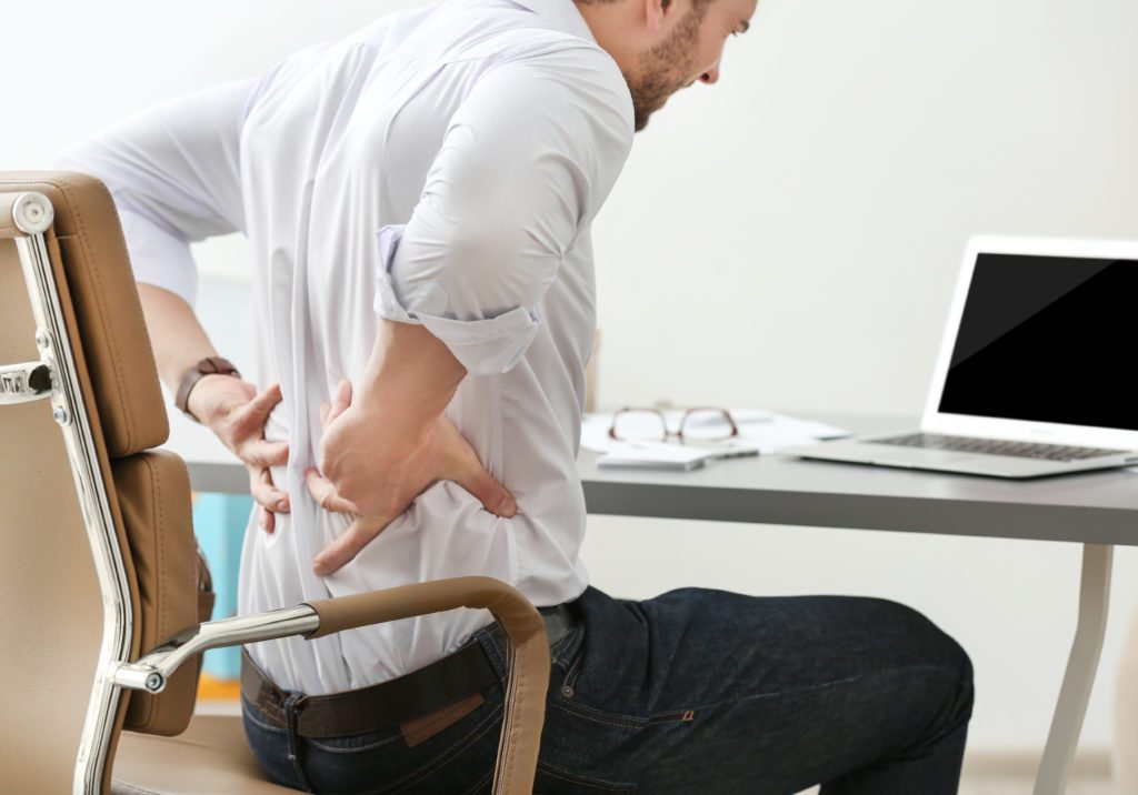 Bilden föreställer en sittande man vid ett skrivbord som håller sig för ryggen